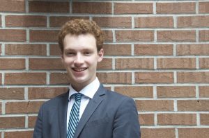 A young man in a suit and blue-striped tie stands in front of a brick wall, smiling at the camera.
