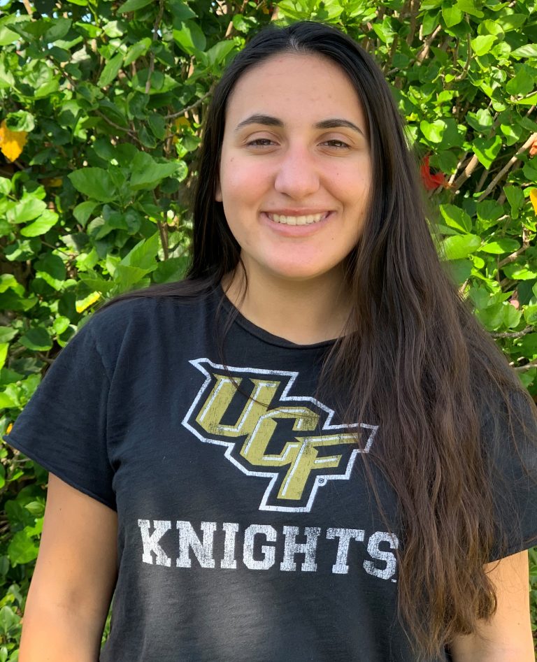 A person with long dark hair is standing outside in front of green foliage, wearing a black UCF Knights T-shirt and smiling at the camera.