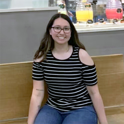 A woman in a black and white striped cold shoulder top and glasses sits on a wooden bench, smiling, with drink dispensers visible in the background.