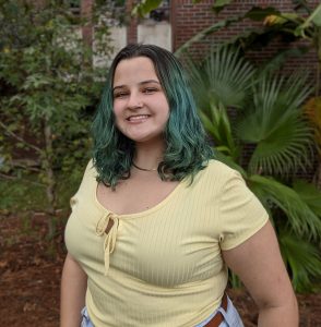 A person with shoulder-length green hair, wearing a light yellow top, stands outdoors in front of greenery and a brick building, smiling at the camera.