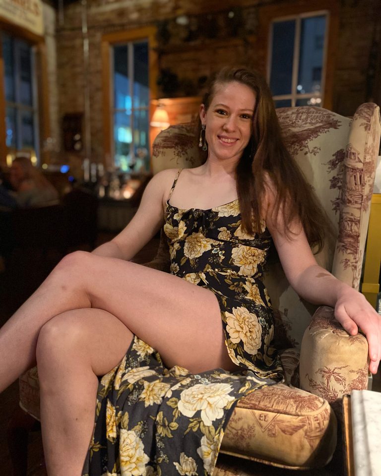 A woman in a floral dress sits comfortably in a patterned armchair in a warmly lit room, smiling at the camera.