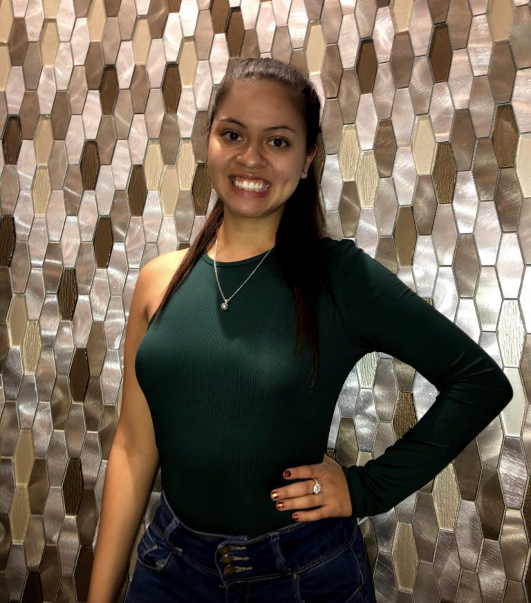 Woman standing and smiling, wearing a dark green one-sleeve top and jeans, with long straight hair, in front of a metallic hexagonal tile wall.
