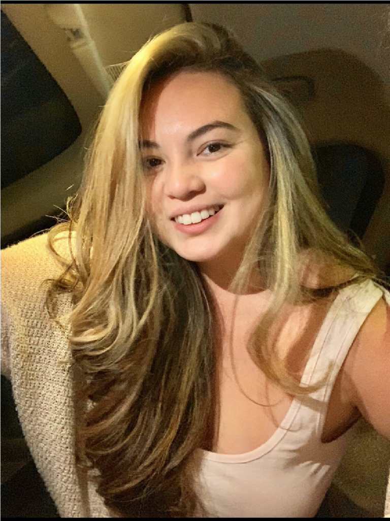 A woman with long, wavy blonde hair and a light-colored top smiles at the camera while sitting indoors.