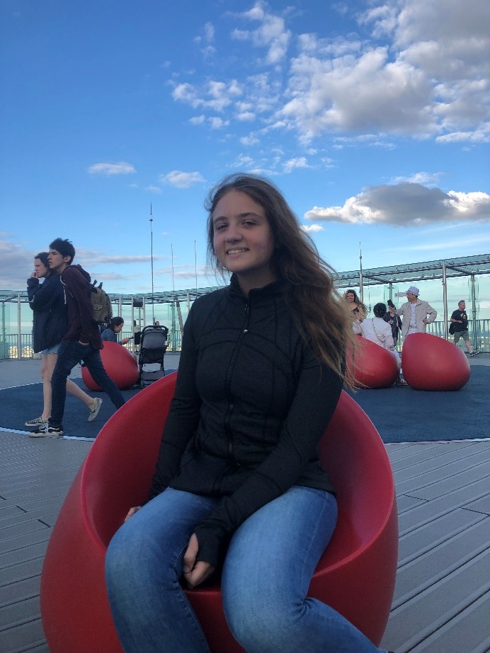 Young woman with long hair and a black jacket sits on a red chair outdoors, with people and a blue sky with clouds in the background.