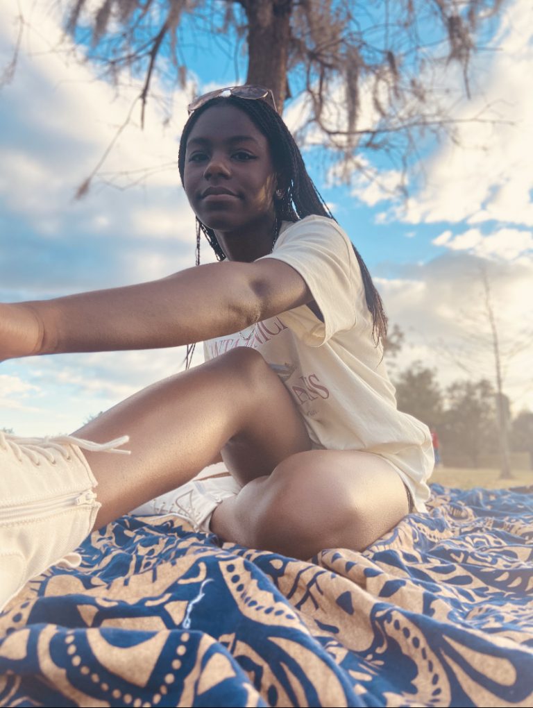 A woman sits on a patterned blanket outdoors, with one knee bent and trees in the background under a partly cloudy sky.