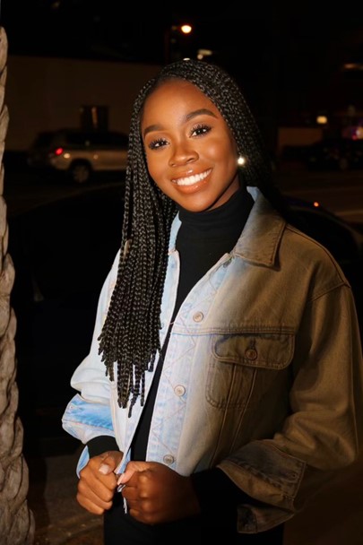 A woman with long braided hair smiles at the camera, wearing a light denim jacket over a black turtleneck, standing outdoors at night.