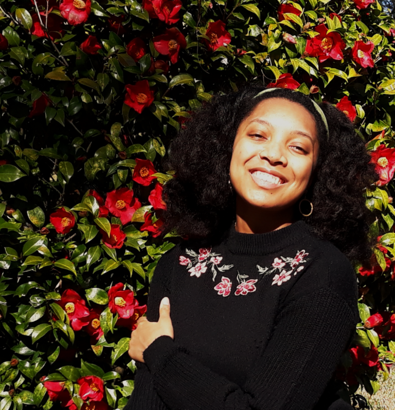 A woman with curly hair smiles in front of a lush bush covered in red flowers, wearing a black sweater with floral embroidery.