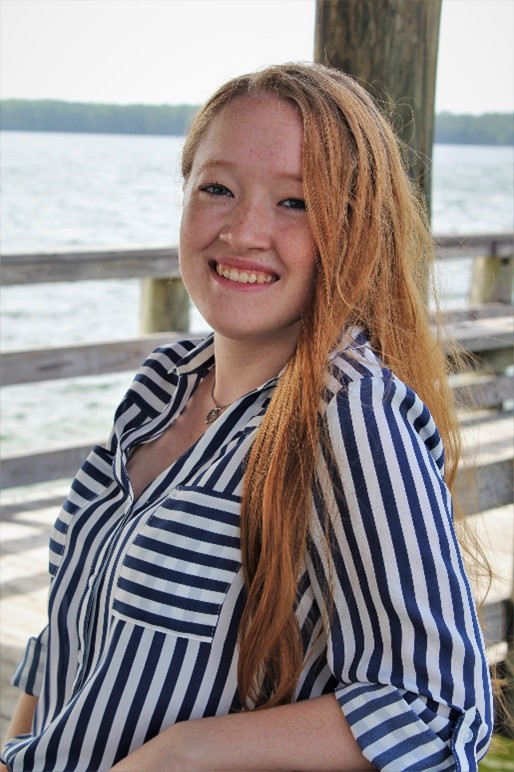 A young woman with long red hair and a striped shirt smiles while standing by a wooden railing near a body of water.