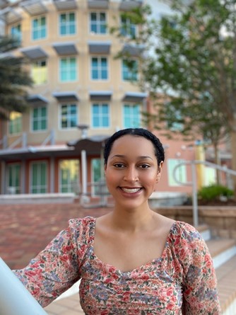 A woman wearing a floral top stands outside in front of a modern building with large windows, smiling at the camera. Trees and stairs are visible in the background.