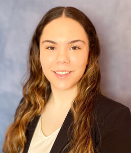 A woman with long wavy brown hair, wearing a black blazer and light-colored top, poses in front of a plain blue background.