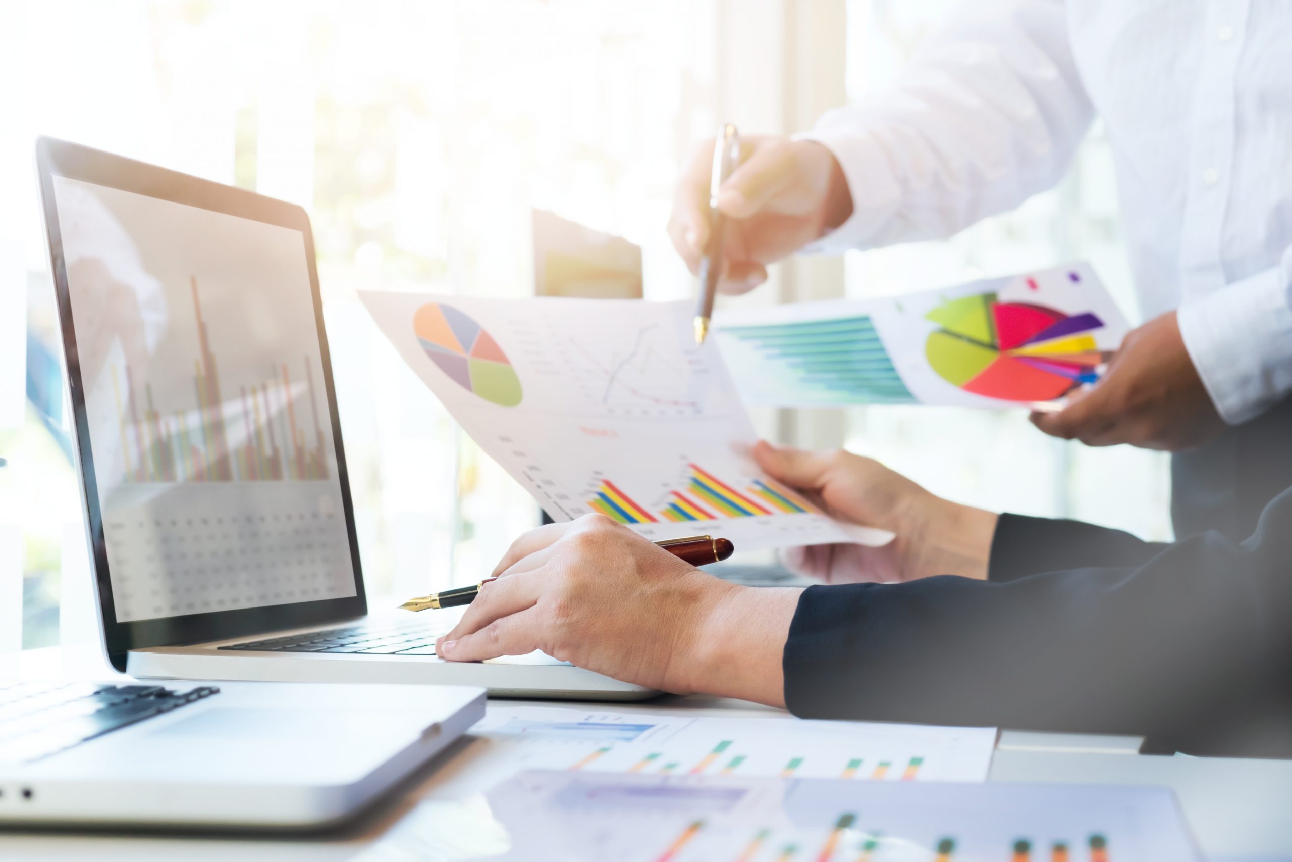 Two people review and discuss printed charts and graphs at a desk with a laptop, analyzing data and financial reports in a brightly lit office setting.