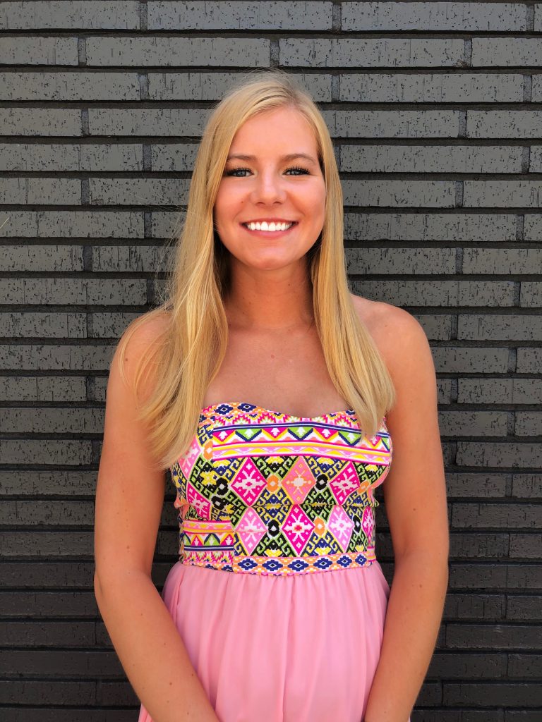 A woman with long blonde hair wearing a strapless, patterned dress stands smiling in front of a dark brick wall.