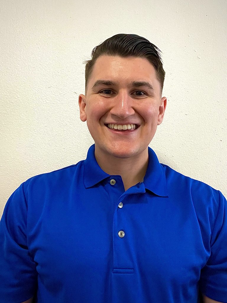 A young man with short dark hair, wearing a bright blue polo shirt, stands smiling in front of a plain white wall.
