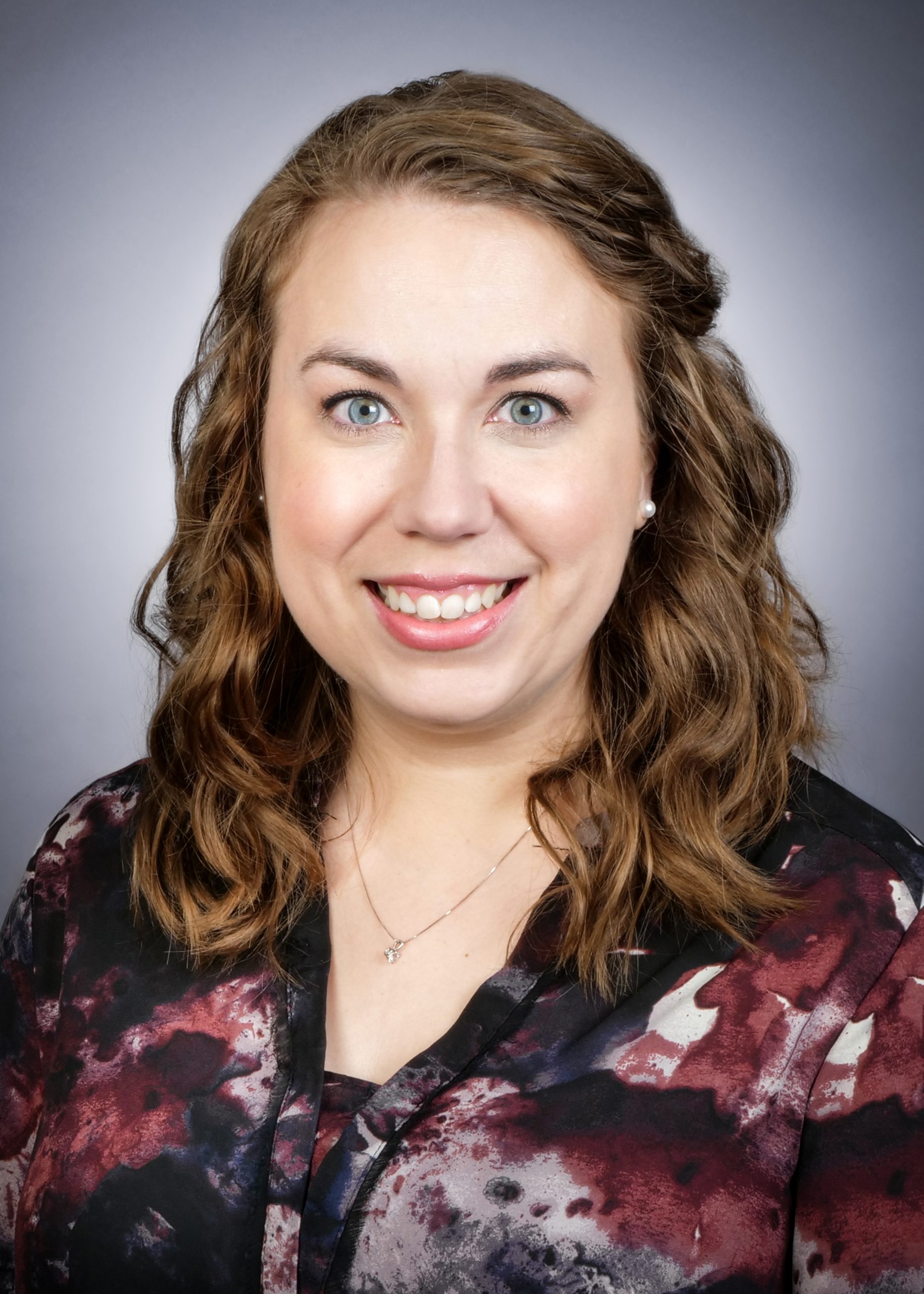 A woman with wavy, shoulder-length brown hair smiles at the camera. She is wearing a patterned blouse and a necklace, posed against a neutral gray background.