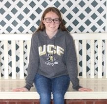 A person wearing glasses and a UCF Knights hoodie sits on a white bench in front of a white lattice background.