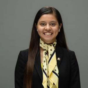 A woman with long dark hair wearing a black blazer and a yellow patterned scarf smiles at the camera against a plain gray background.