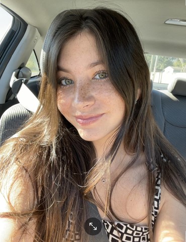 Young woman with long brown hair and freckles sits in a car seat, wearing a patterned top and seatbelt, smiling at the camera in natural sunlight.