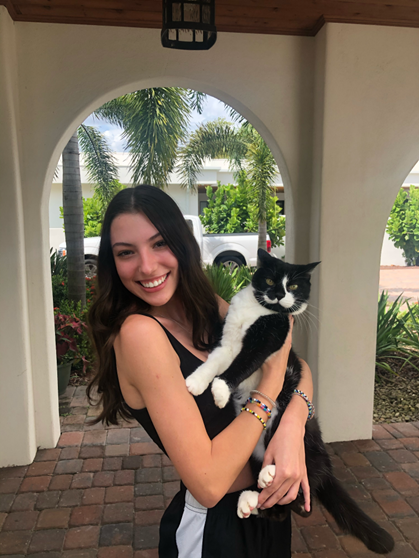 A young woman stands outside under an archway, smiling and holding a black and white cat in her arms.