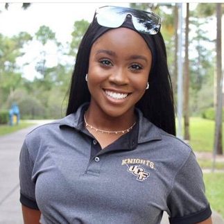 A woman wearing a UCF Knights polo shirt and sunglasses on her head smiles outdoors on a pathway lined with trees.