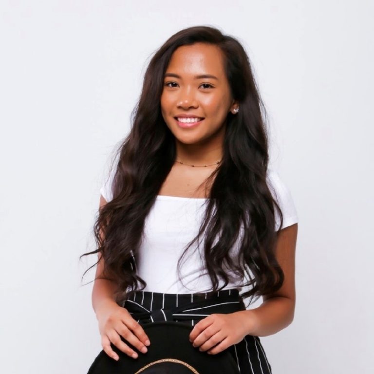 A young woman with long wavy hair wearing a white top and black striped pants stands against a plain white background, smiling at the camera.