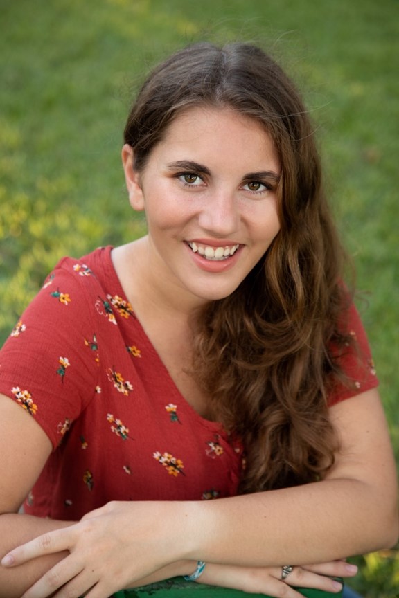 A woman with long brown hair wearing a red floral top sits outdoors on grass, smiling at the camera.