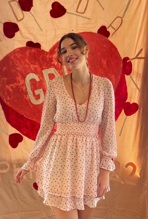 A woman in a light pink, polka-dotted dress stands in front of a beige backdrop with large red hearts and the letters "GRD" painted on it.