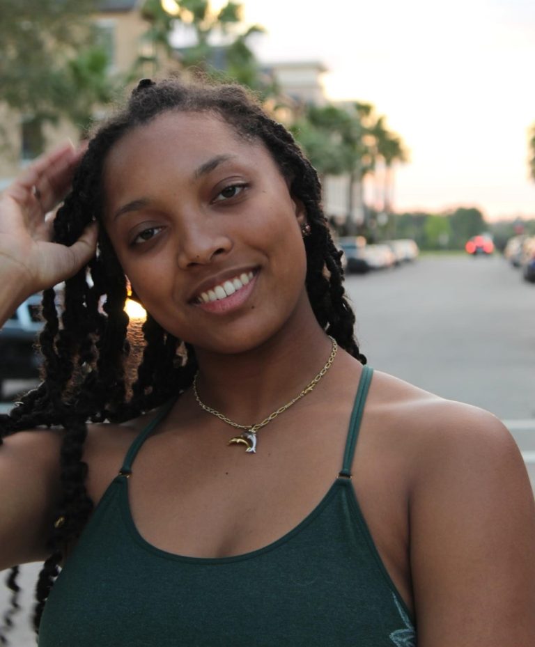 A woman with long braided hair smiles at the camera, wearing a dark green tank top and a dolphin necklace, standing on a street lined with palm trees at sunset.