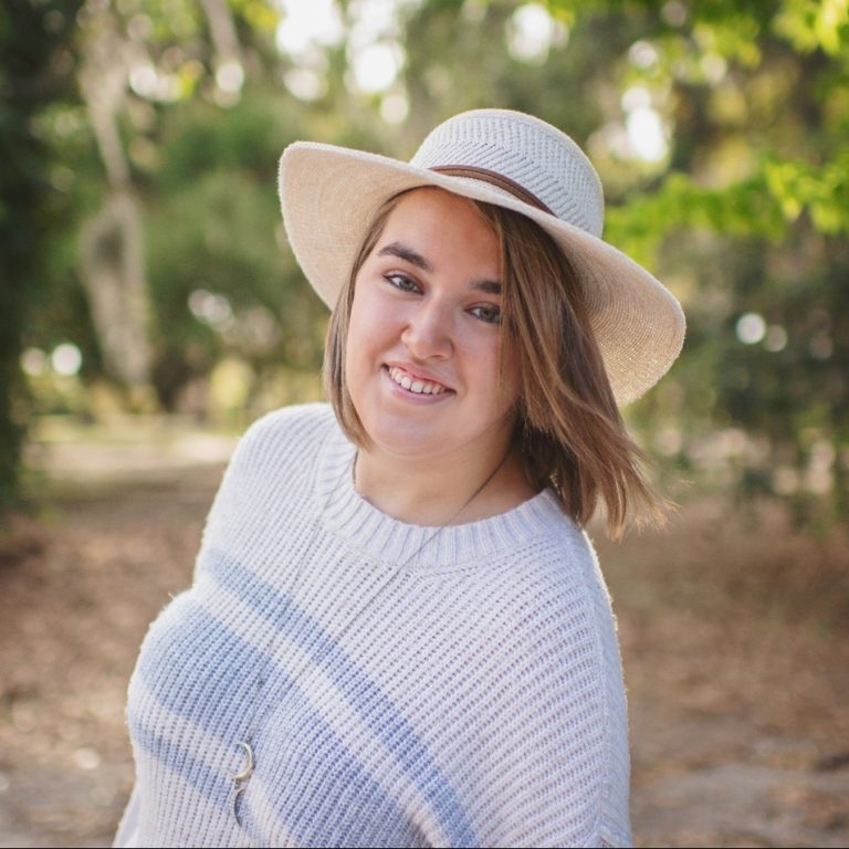 A woman wearing a light-colored sweater and a wide-brimmed hat smiles outdoors with trees in the background.