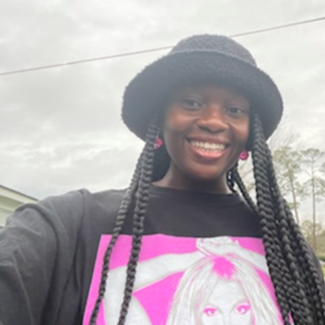 A person with long braids wearing a black bucket hat and a black T-shirt with a pink graphic smiles outdoors under a cloudy sky.
