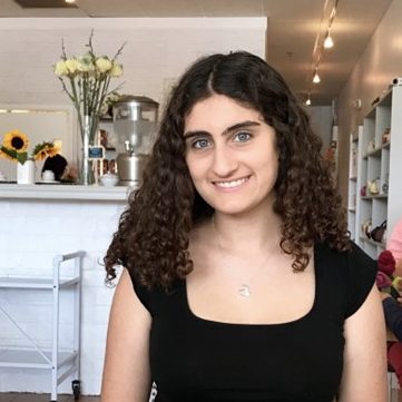 A woman with curly brown hair and a black top sits and smiles in a cafe with flowers and shelves in the background.