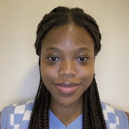 A woman with long braided hair and a blue sweater vest smiles slightly while facing the camera against a plain light-colored background.