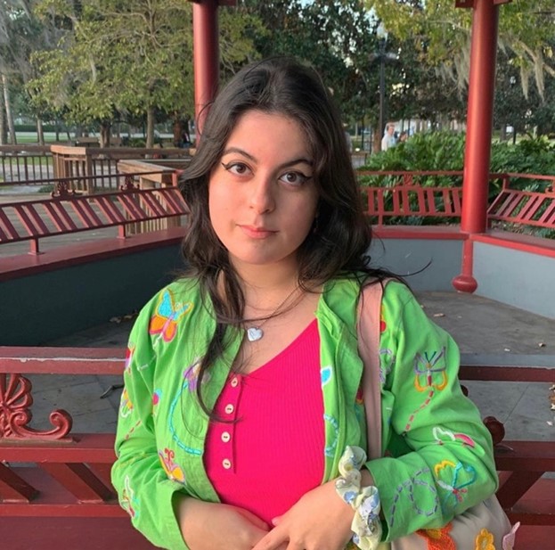 A young woman with dark hair stands in front of a red gazebo, wearing a green jacket with colorful designs over a pink top, looking at the camera. Trees and benches are in the background.