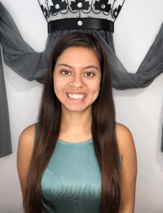 A young woman with long brown hair, wearing a sleeveless teal top, smiles in front of a wall with draped gray fabric and a decorative black-and-white floral element.