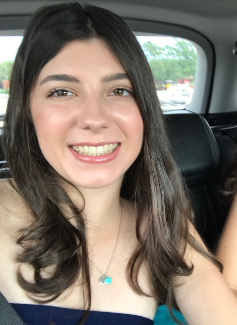 A young woman with long brown hair and a necklace smiles while sitting in the backseat of a car.
