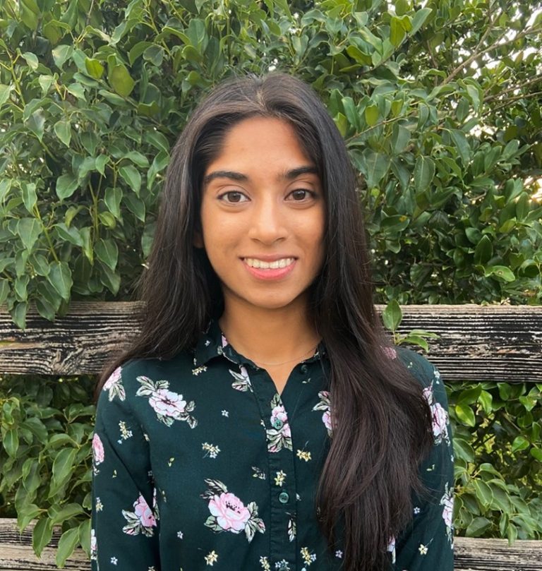 A young woman with long dark hair, wearing a dark floral blouse, stands in front of a wooden fence and green leafy bushes, smiling at the camera.