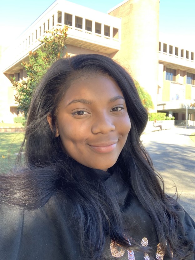 A woman with long dark hair smiles outdoors in front of a large tan brick building on a sunny day.