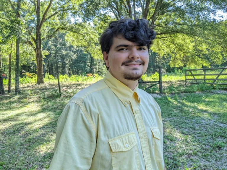 A person with short dark hair and a mustache, wearing a yellow button-up shirt, stands outdoors in a sunlit grassy area with trees and a wooden fence in the background.
