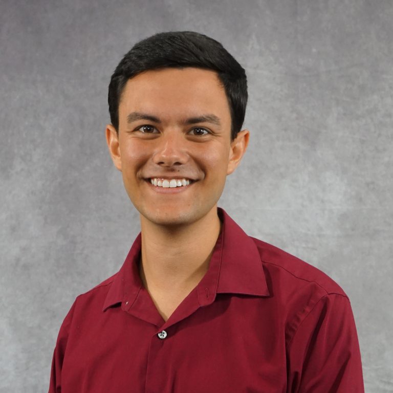 A young man with short dark hair wearing a maroon collared shirt smiles at the camera in front of a gray background.