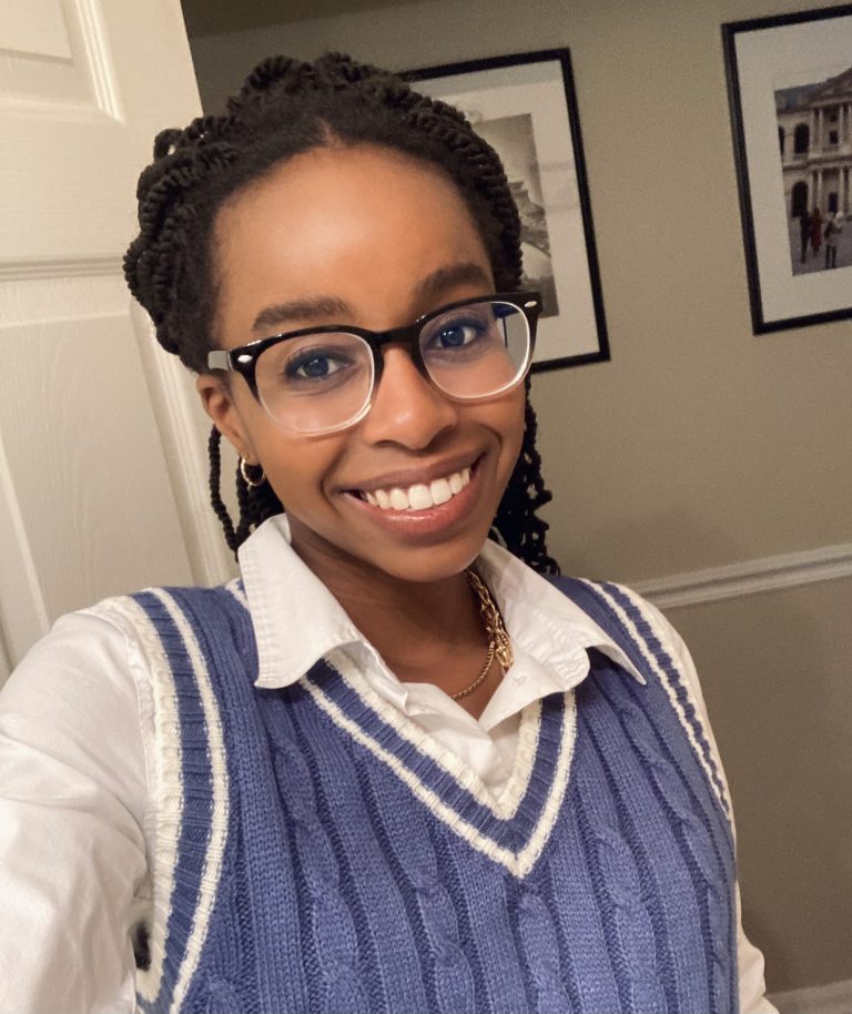 A woman wearing glasses, a white shirt, and a blue sweater vest smiles at the camera; framed pictures hang on the beige wall behind her.