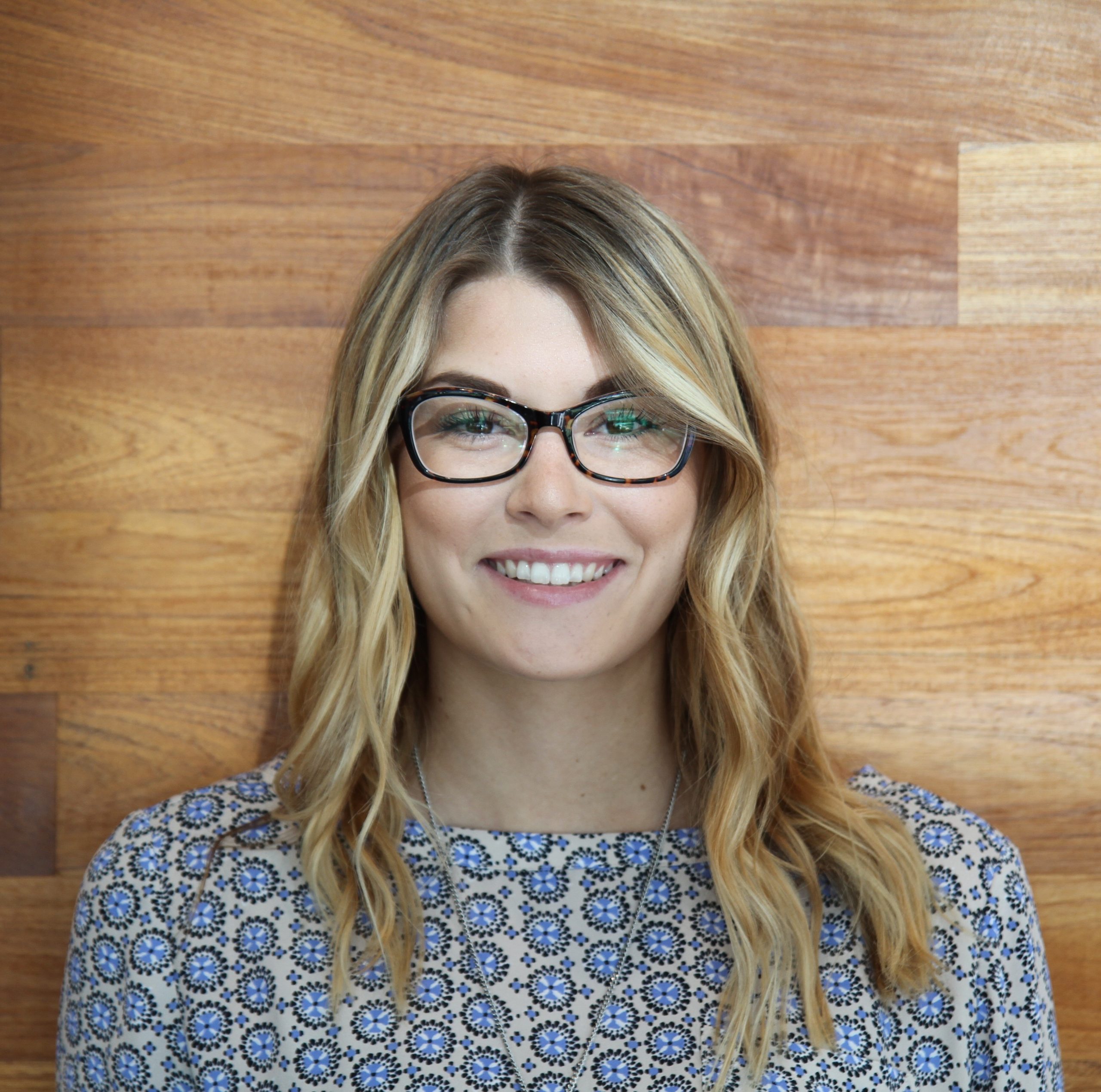 A woman with blonde hair and glasses smiles in front of a wooden wall, wearing a patterned blue and white top.