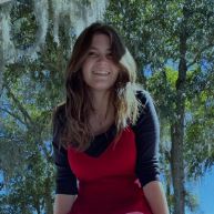 A woman with long brown hair, wearing a red dress over a black shirt, smiles while standing outdoors with trees and sunlight in the background.