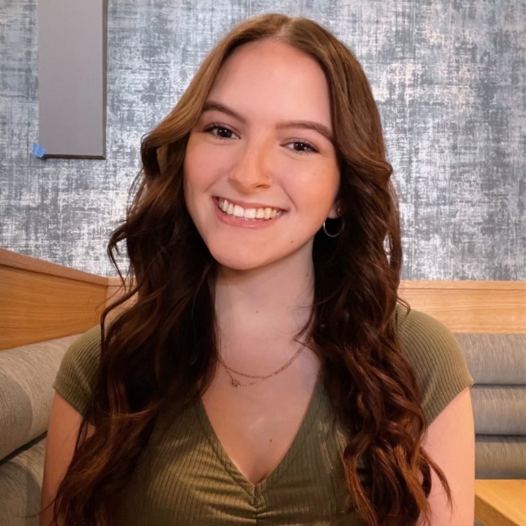 A young woman with long wavy brown hair, wearing a green top, sits and smiles at the camera in a modern indoor setting with a textured wall and cushioned seating.