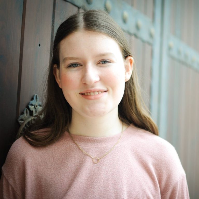 A young woman with long brown hair, wearing a pink top and a gold necklace, stands in front of a wooden door with decorative metal accents, smiling at the camera.