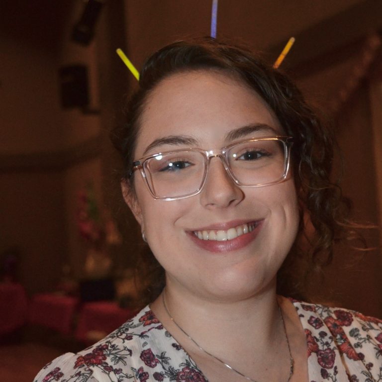 A woman with glasses and curly hair smiles at the camera, wearing a floral top and colorful glow sticks in her hair.