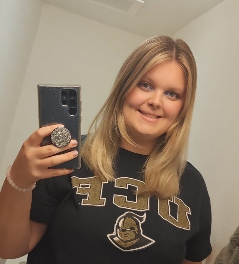 A woman with straight blonde hair takes a mirror selfie, wearing a black UCF t-shirt and smiling.