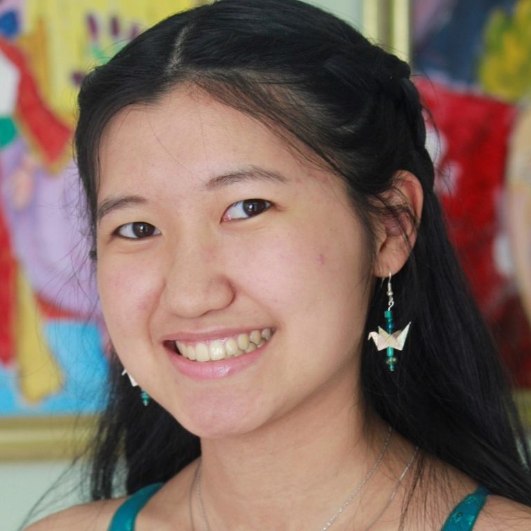 A young woman with long dark hair smiles at the camera, wearing origami crane earrings and a necklace, with colorful artwork in the background.