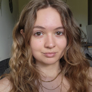 A young woman with wavy light brown hair and a neutral expression looks at the camera. She is wearing layered gold necklaces and sits indoors with a blurred background.