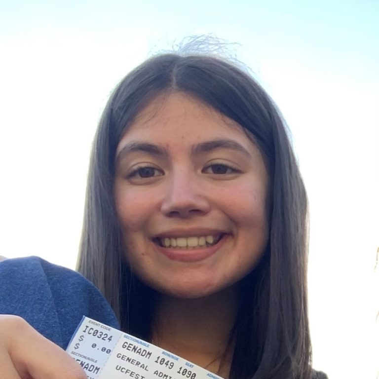 A young woman with long dark hair smiles at the camera while holding up a ticket with both hands. The sky is visible in the background.