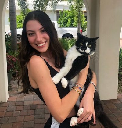 A woman with long brown hair smiles while holding a black and white cat outdoors on a brick patio with greenery in the background.