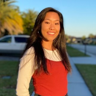Woman with long dark hair stands outside on a sunny day, wearing a red top over a white shirt, with a street and greenery in the background.